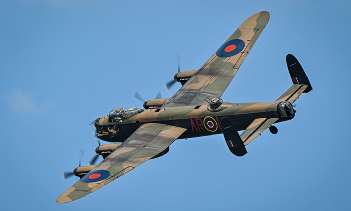 A WWII-era Avro Lancaster bomber in brown-green camouflage, RAF roundels on its wings, banking left in clear blue sky with propellers blurred in motion.