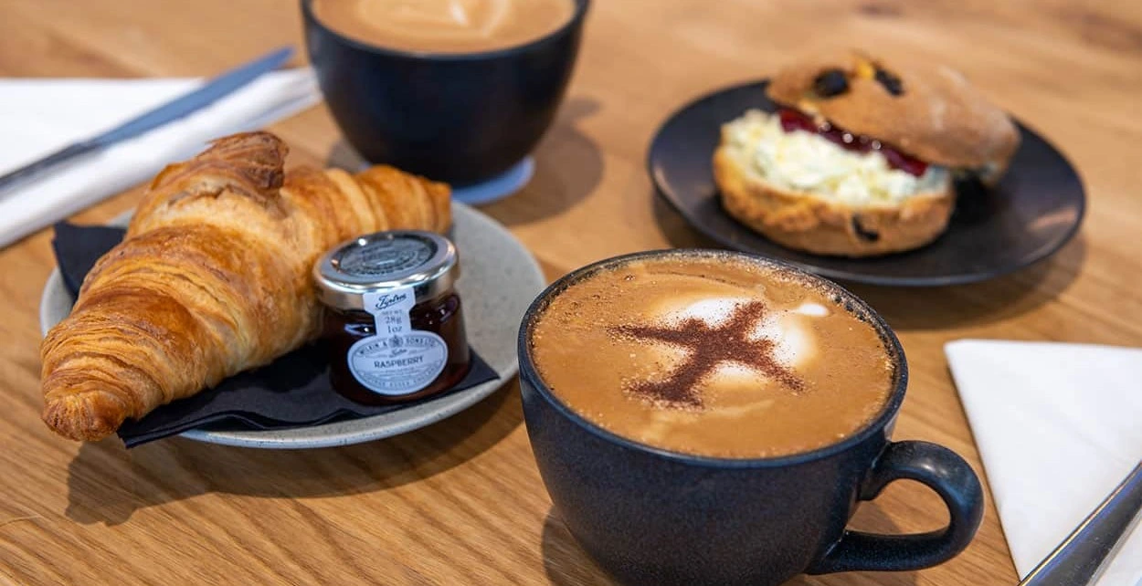 A round wooden table with two lattes, a croissant with jam and butter, a scone with cream and jam, and a menu standing upright. One coffee has a chocolate aeroplane design on the froth.
