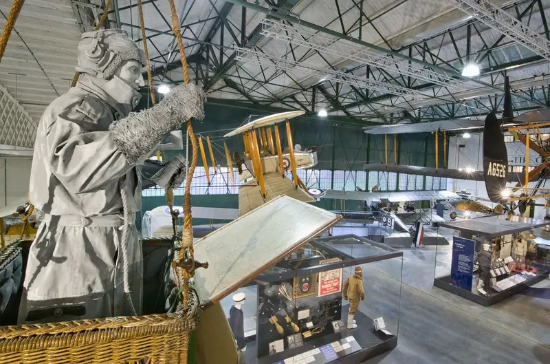 A museum scene featuring a pilot mannequin in a wicker basket, aircrafts and exhibits in an industrial hall.