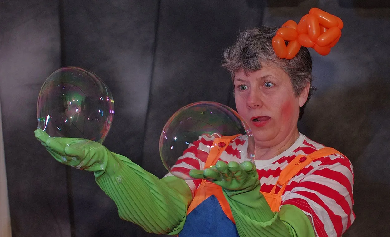 Performer in colourful costume with green gloves holds two large soap bubbles during a science show