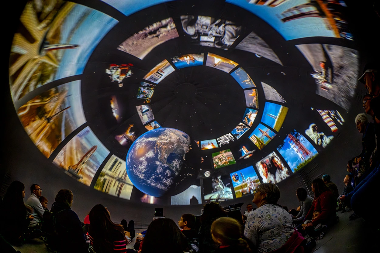 A domed theater displays projected images of Earth and space in a spiral pattern, surrounded by an audience gazing upward.