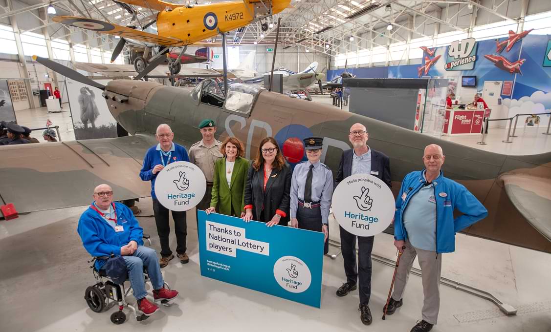 A group of people pose in front of a vintage military aircraft indoors, holding Heritage Fund and National Lottery signs in the Museum.